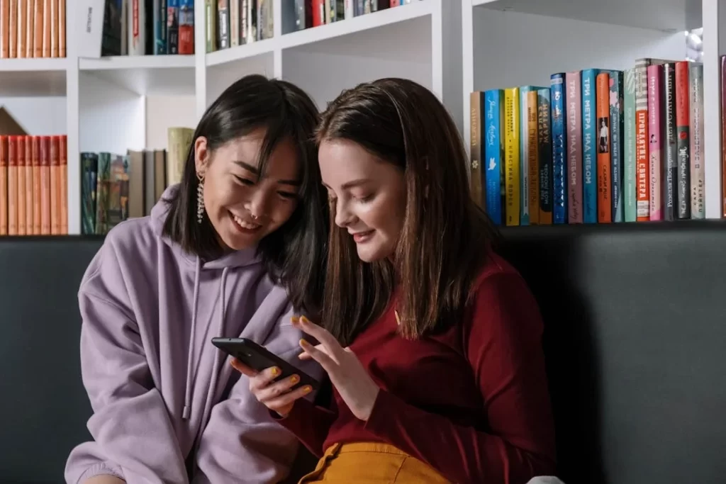 Dua remaja perempuan sedang tersenyum sambil melihat sesuatu di ponsel mereka di depan rak buku.